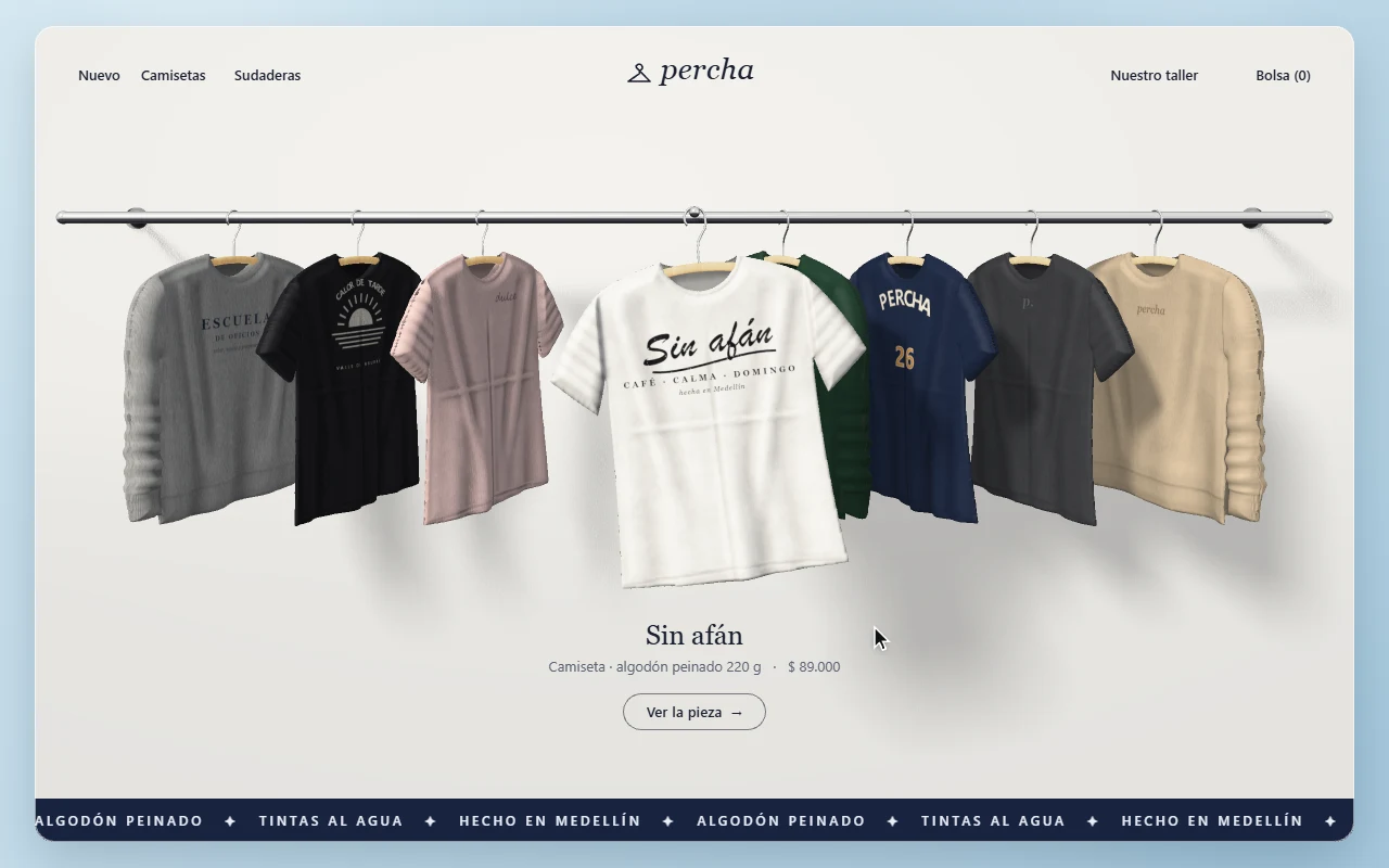 Página PERCHA: ocho camisetas y sudaderas cuelgan de perchas de madera en una barra cromada, giradas de perfil hacia el centro; al frente, una camiseta blanca con la frase «Sin afán» en letra caligráfica, con pliegues y sombras suaves sobre la pared.